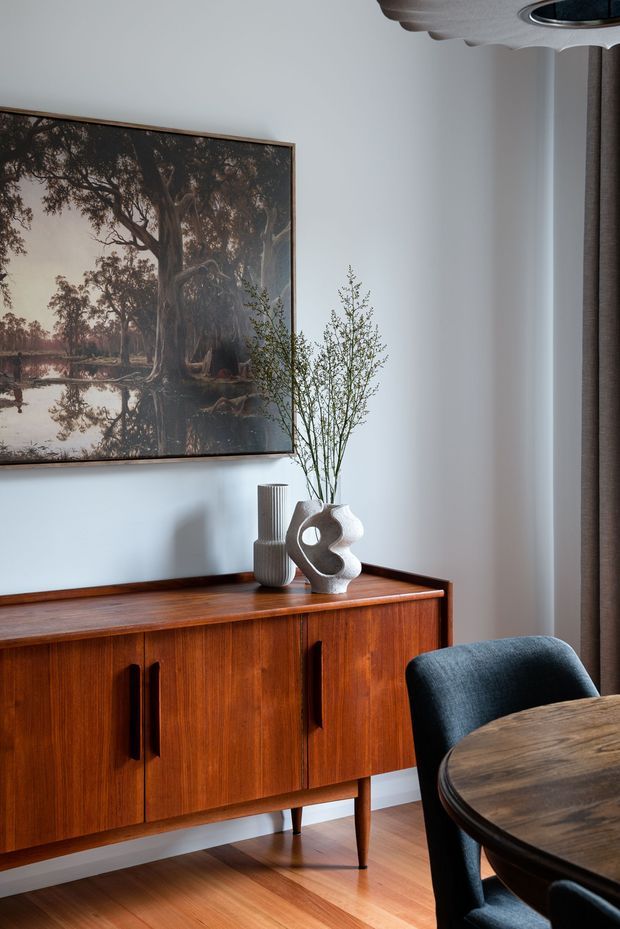 A refined dining area featuring a mid-century timber sideboard with tapered legs, a round timber dining table, and dark upholstered dining chairs. The room has pale walls, warm timber flooring and joinery, a large framed landscape artwork, sculptural ceramic vases, and soft natural light from a curtained window.
