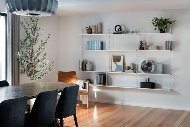 Bright, refined dining and living area with a large timber dining table, dark upholstered dining chairs, a woven accent chair, and extensive open white shelving displaying books and decorative objects. The room has pale walls, timber flooring, indoor greenery, large window glazing, and a sculptural contemporary pendant light.