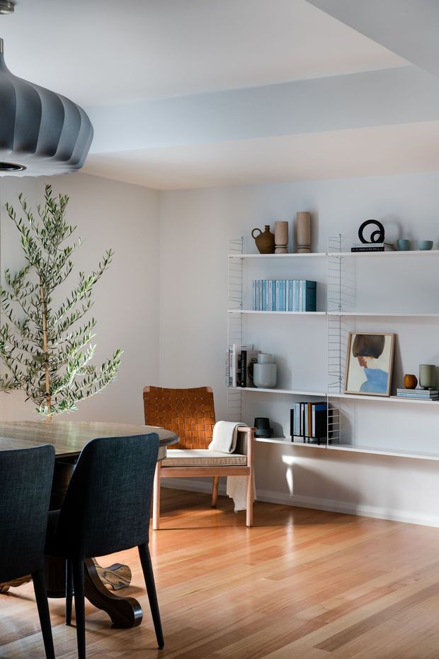 A bright, refined open-plan dining and living area with white walls, warm timber flooring, a dark sculptural pendant light, and a built-in white wire-frame shelving system displaying ceramics, books, and artwork. A timber dining table with dark upholstered chairs sits beside a woven leather lounge chair and an indoor olive tree. Natural light creates a calm, contemporary atmosphere.