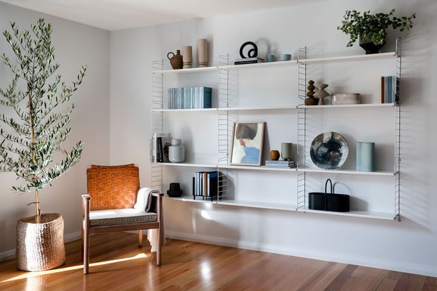 Bright, curated living room featuring a large wall-mounted open shelving system filled with books, ceramics, artwork and decorative objects. A woven lounge chair, tall potted tree, and warm timber flooring add texture and warmth against pale grey-white walls.