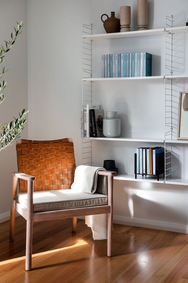 A bright, warmly styled reading nook with a cognac leather woven lounge chair, light upholstered seat cushion, and draped knitted throw. A white wall-mounted wire shelving system displays books, ceramic vessels, and decorative objects. Timber flooring, soft white walls, and strong natural sunlight create a calm mid-century-inspired interior.