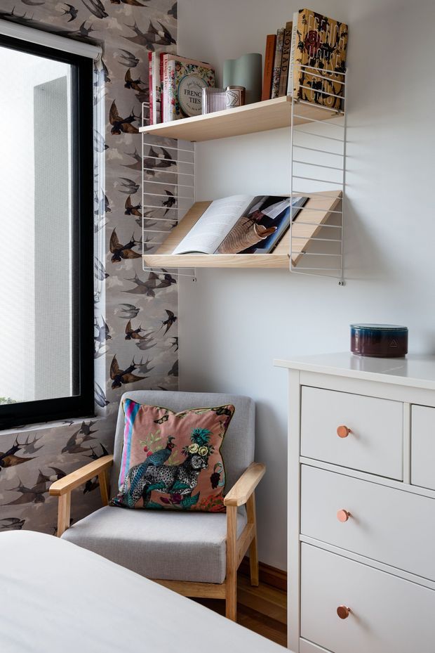 A compact, thoughtfully styled bedroom corner with a grey upholstered timber armchair, colourful animal-print cushion, white chest of drawers, and wall-mounted open shelving displaying books and magazines. A large black-framed window is dressed with bird-patterned wallpaper or fabric, while timber flooring and neutral walls create a warm backdrop.