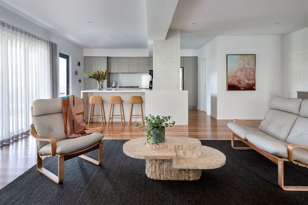 Bright open-plan contemporary living room with a kitchen visible beyond. The space features white walls and cabinetry, warm timber flooring, a large dark area rug, pale upholstered lounge furniture, a sculptural round timber coffee table, and floor-to-ceiling sheer curtains. A central white structural wall or column partially separates the living and kitchen areas.
