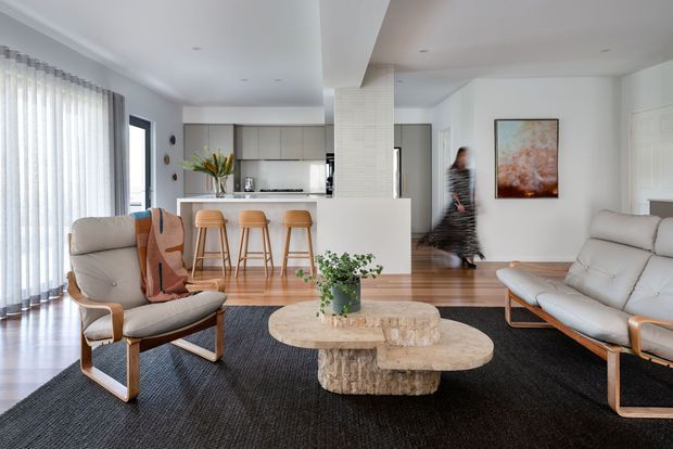 A bright, open-plan contemporary living area with a white kitchen and peninsula breakfast bar in the background. The room features pale timber flooring, a large dark textured rug, sculptural stone coffee tables, light upholstered lounge chairs and sofa, sheer curtains, and neutral white cabinetry. A blurred figure is moving through the space, with artwork and indoor plants adding colour.