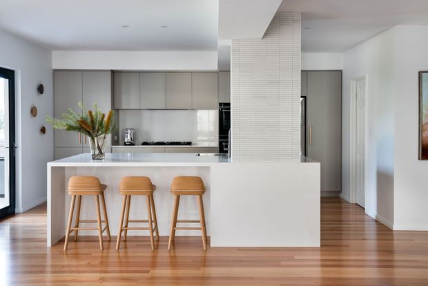 Bright open-plan contemporary kitchen with a large white island, three timber bar stools, grey flat-panel cabinetry, stainless steel appliances, and a white tiled feature column. Warm timber flooring extends into the adjacent hallway and living area.