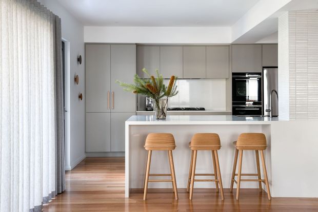 Bright contemporary kitchen with full-height matte grey cabinetry, a white island and benchtop, integrated black appliances, timber flooring, and three timber bar stools. Sheer vertical blinds and subtle decorative styling contribute to a clean, refined residential interior.