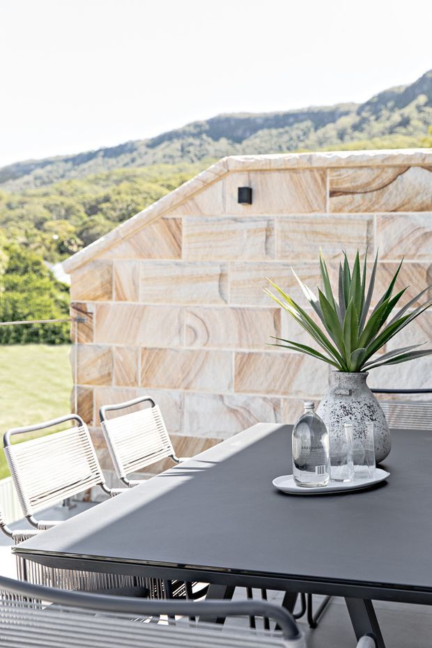 A bright outdoor dining terrace with a large dark rectangular table and woven sling chairs. The terrace is enclosed by a pale natural-stone block wall with a sloped parapet detail, and a potted architectural plant sits on a tray at the centre of the table. A lush mountainous landscape is visible beyond the terrace.