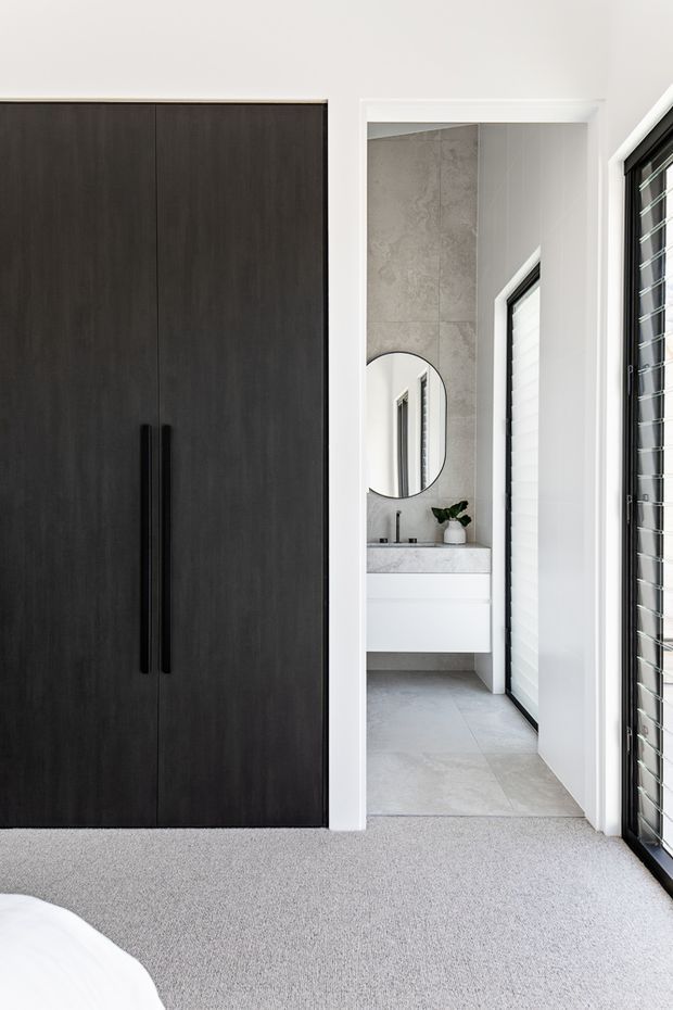 A bright, contemporary bedroom looking toward an adjoining bathroom. The bedroom features a full-height dark timber wardrobe with long black handles, light grey carpet, and a partial white bed in the foreground. Through the open doorway is a compact bathroom with pale grey large-format tiled walls and floor, a floating white vanity, round mirror, black tapware, and a narrow window with white shutters. Black-framed glazing with horizontal louvres is visible along the right side.