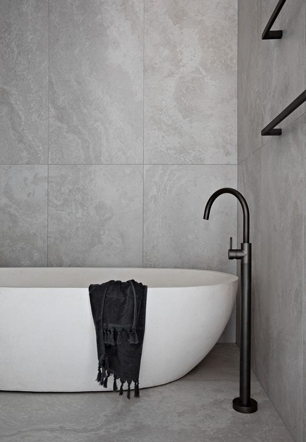 A minimalist bathroom featuring a freestanding white oval bathtub, large-format grey stone-look wall and floor tiles, and a matte black floor-mounted curved bath mixer. Minimal black wall-mounted fixtures and a dark towel provide contrast against the restrained grey and white palette.