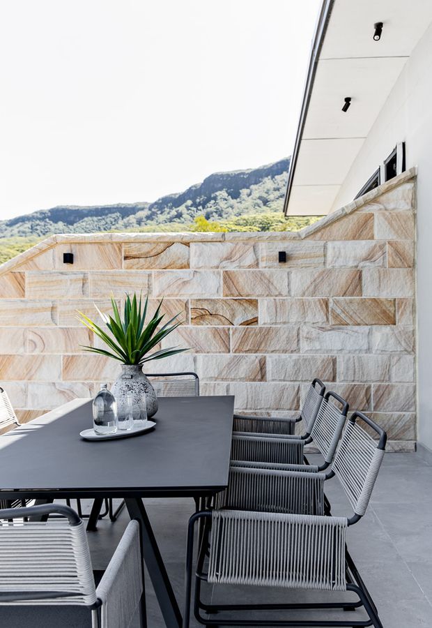 A contemporary outdoor dining terrace with a long dark rectangular table and rope-woven dining chairs, enclosed by a warm-toned natural stone feature wall. The terrace has grey tiled flooring, a white soffit with black recessed or surface-mounted lights, and views toward a green mountain landscape. A potted spiky plant and table setting add a restrained decorative touch.