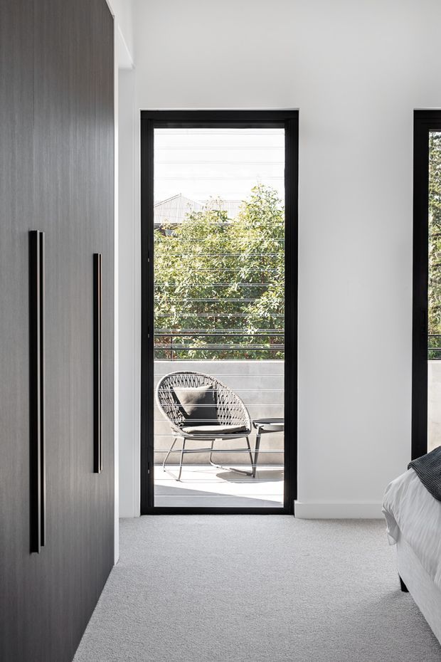 Bright contemporary bedroom interior with a soft grey carpet, white walls, a large dark built-in wardrobe with long vertical handles, and a black-framed glazed door opening onto a small balcony or terrace. A partial bed is visible at right, while the balcony contains a woven lounge chair and overlooks leafy surroundings.