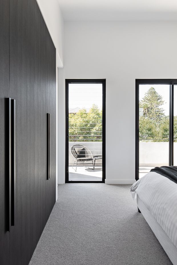Bright contemporary bedroom with a large dark timber built-in wardrobe along the left wall, soft grey carpet, white walls and ceiling, and black-framed glazing opening onto a balcony or terrace. A partially visible bed occupies the right foreground, with outdoor seating and leafy views beyond the doors.
