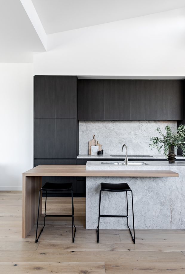 A refined contemporary kitchen with dark timber cabinetry, a large pale stone island with integrated seating, and light oak flooring. The minimalist space features white walls and ceiling, a timber breakfast bar overhang, black bar stools, a stainless-steel tap and sink, and understated styling with greenery.