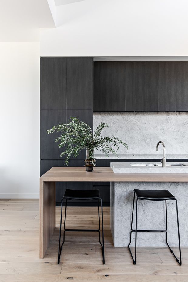 A refined contemporary kitchen featuring dark timber cabinetry, a pale stone or concrete-textured island with integrated sink, and a timber breakfast bar supported by black bar stools. Light timber flooring, white walls, and minimal styling create a clean, restrained interior.