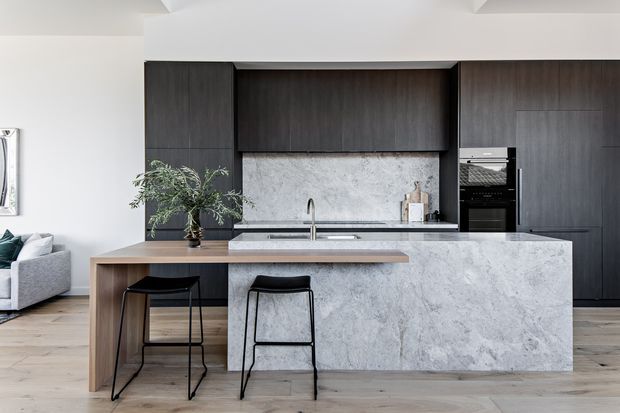 Contemporary open-plan kitchen with a large marble-look island, integrated dark timber cabinetry, a stone splashback, and two black bar stools. Light timber flooring and white walls create a clean, refined contrast, with a living area partially visible at left.