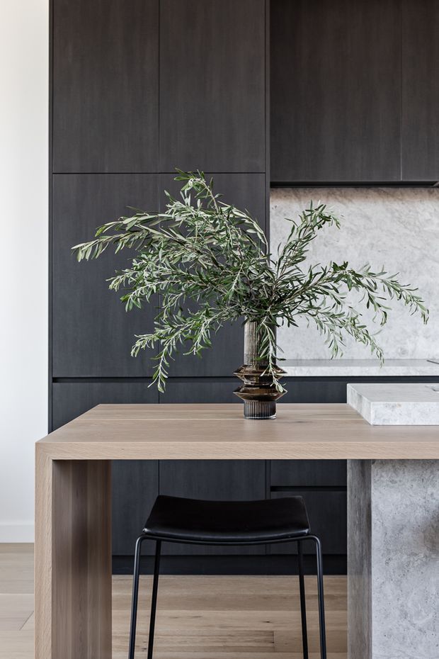 A refined contemporary kitchen and dining area featuring dark charcoal full-height cabinetry, a pale natural timber dining table, a black chair, and a pale grey stone island or benchtop. A sculptural leafy branch in a dark vase forms the central styling element.