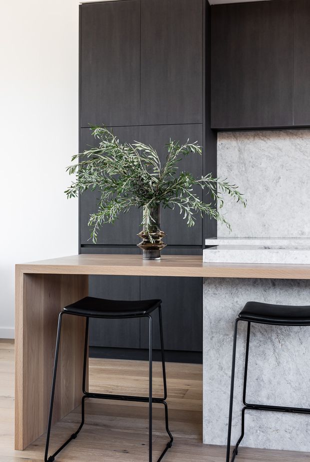 A refined contemporary kitchen featuring tall dark timber cabinetry, a pale stone waterfall island and raised stone splashback, and a light timber breakfast counter with black bar stools. The restrained palette is accented by an olive branch arrangement and warm timber flooring.