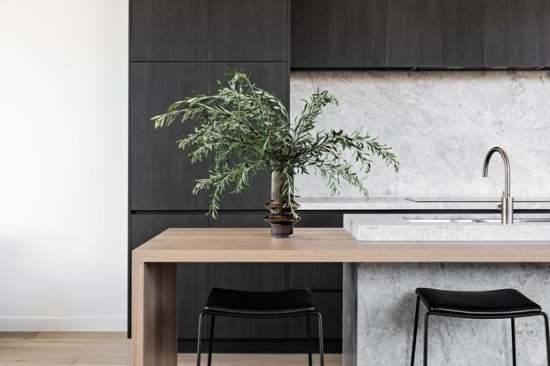 Contemporary kitchen featuring dark timber cabinetry, a pale stone or concrete-textured splashback, a light timber island with a substantial pale benchtop, and black bar stools. A sculptural branch arrangement adds an organic accent to the restrained, monochromatic scheme.