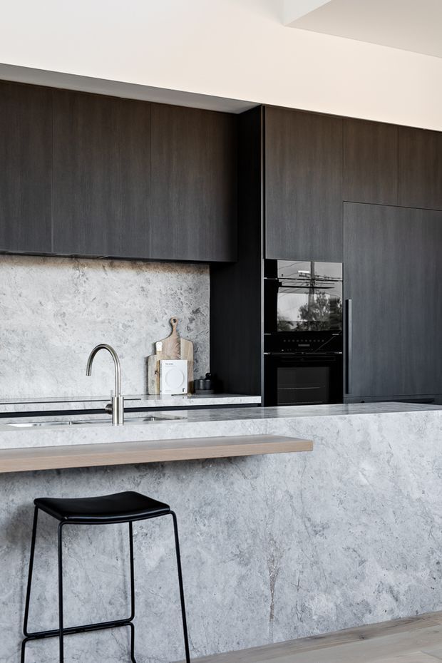 Contemporary kitchen with dark timber cabinetry, integrated black appliances, a large pale stone island and matching stone splashback. A slim timber breakfast ledge and black bar stool add contrast, while stainless steel tapware and minimal styling create a refined, monochromatic look.