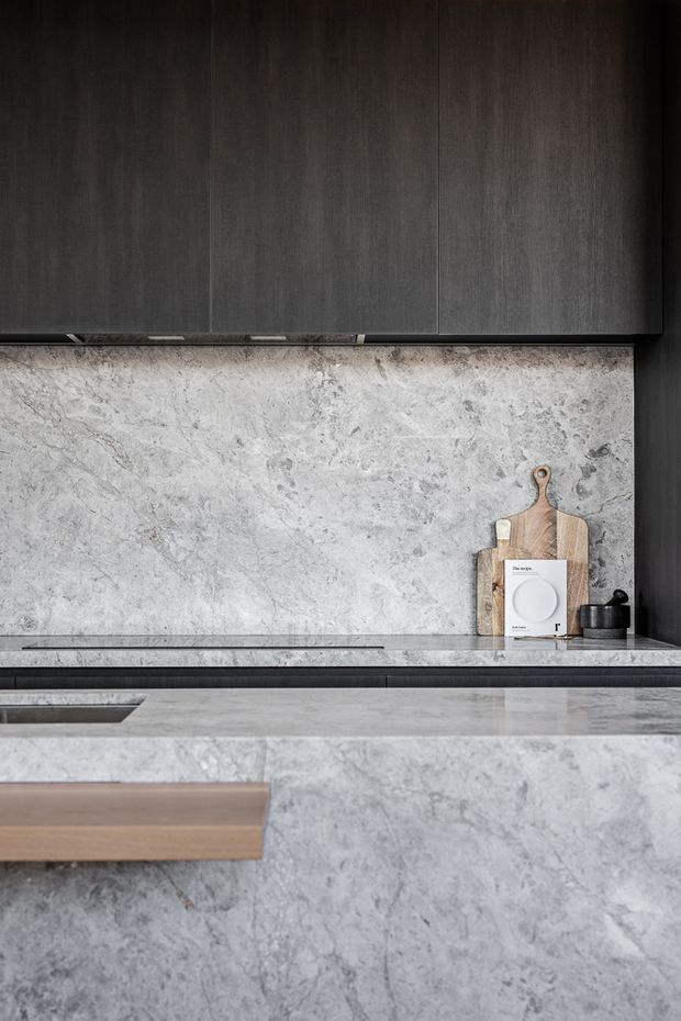Close-up view of a refined contemporary kitchen with dark vertical-grain cabinetry, a pale grey heavily veined stone splashback and matching island surfaces. A timber cutting board and small black countertop accessories provide restrained contrast. The composition emphasizes the material palette and integrated joinery rather than the full room layout.