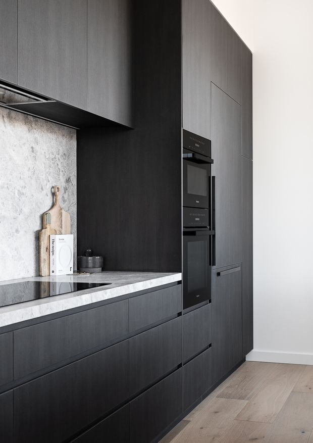 A refined contemporary kitchen featuring tall dark charcoal cabinetry, integrated black appliances, a pale veined stone benchtop and splashback, and light natural timber flooring. The cabinetry extends floor to ceiling, creating a streamlined built-in wall with minimal visible hardware.