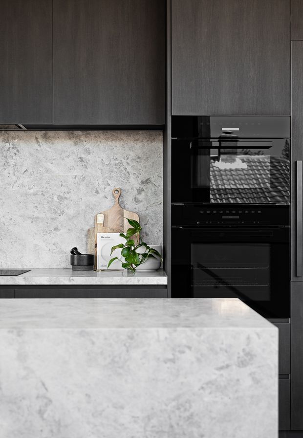 Close-up view of a refined contemporary kitchen featuring dark charcoal cabinetry, integrated black appliances, and pale grey veined stone used across the splashback, benchtops, and island. A timber chopping board and potted plant provide small natural accents.