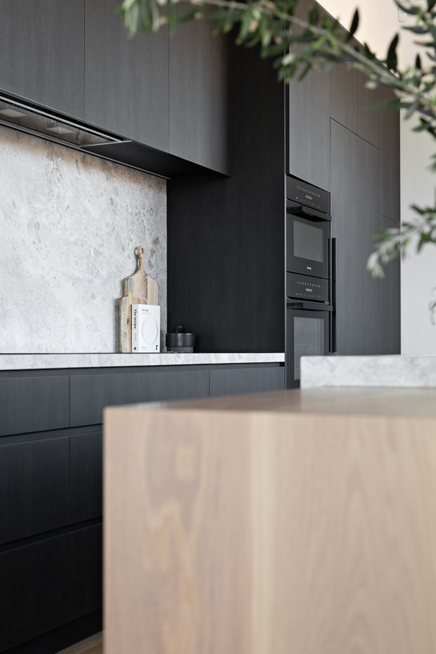 A refined contemporary kitchen featuring tall matte dark cabinetry, integrated black appliances, a pale textured stone splashback, and a light timber island with a pale stone benchtop. Minimal styling and soft greenery create a warm, sophisticated contrast against the dark joinery.