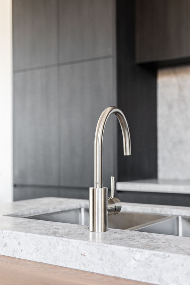 Close-up of a contemporary kitchen sink area featuring a tall curved stainless-steel tap mounted in a pale stone benchtop. A double-bowl stainless-steel sink, dark cabinetry, grey tiled wall surfaces, and additional pale stone counters are visible in the background.