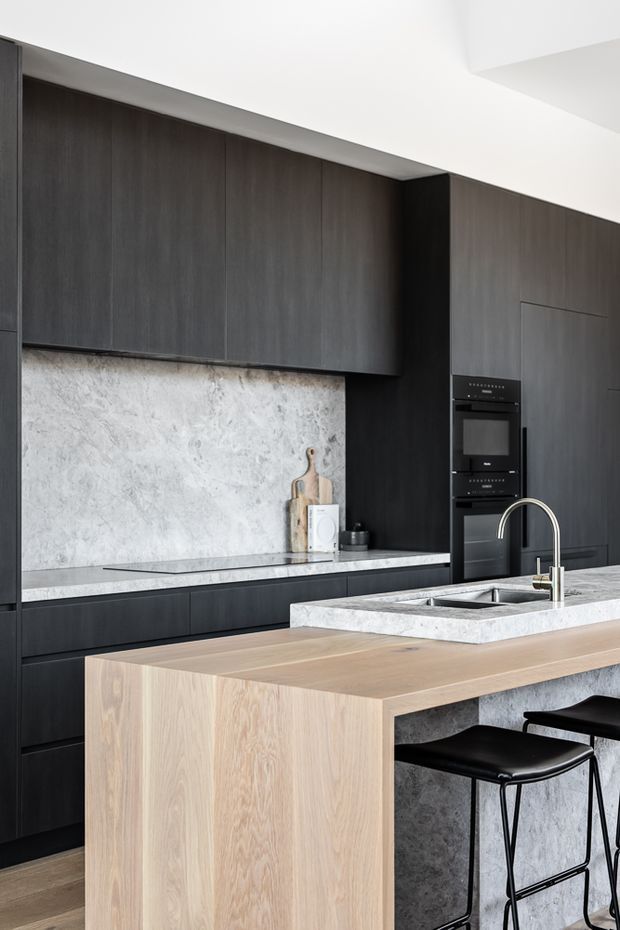 A contemporary kitchen with floor-to-ceiling dark wood cabinetry, integrated appliances, a pale stone backsplash and benchtop, and a large light timber island with a waterfall-style side panel. Black bar stools sit beneath the island, while a stainless steel gooseneck tap and undermount sink occupy the stone work surface. The space has a restrained, high-contrast palette and clean-lined built-in joinery.