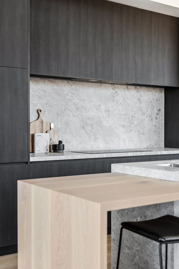 A refined contemporary kitchen featuring dark charcoal cabinetry, a pale marbled stone splashback and benchtop, and a light timber kitchen island with black bar seating. The composition highlights clean, handleless joinery and contrasting natural materials.