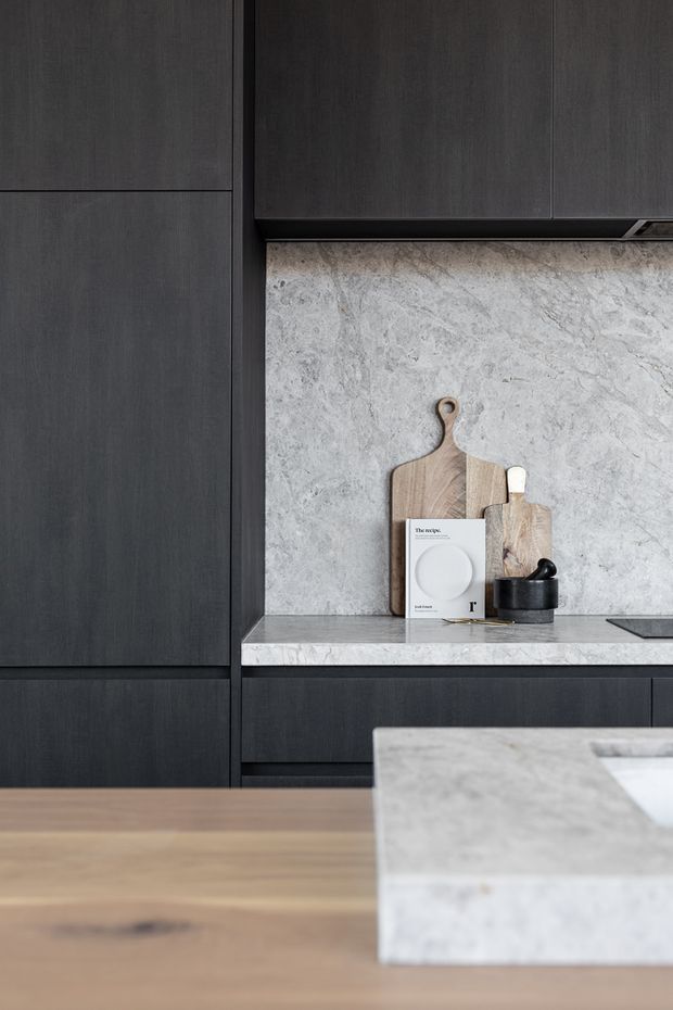 Close-up view of a refined contemporary kitchen featuring charcoal-black built-in cabinetry, a pale veined stone benchtop and matching splashback, and a timber island or dining surface in the foreground. Wooden chopping boards and a small dark vessel provide understated styling accents.
