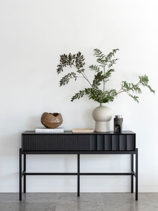 A minimalist interior vignette featuring a slim black console table with vertically fluted cabinet fronts and an open metal frame. The console is styled against a plain white wall with a sculptural off-white vase holding leafy branches, a decorative bowl, books, and a small dark vessel. The floor is a pale grey polished concrete or stone-like finish.