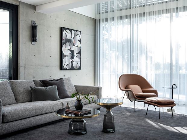A refined contemporary living room with a large grey sectional sofa, sculptural black-and-glass coffee tables, a brown leather lounge chair and ottoman, and sheer full-height curtains. The room features an exposed concrete feature wall, black-framed glazing, minimalist abstract wall art, and abundant filtered daylight.