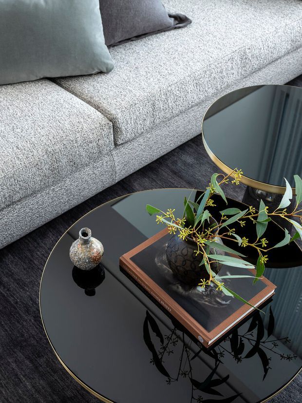 Close-up interior vignette of a contemporary living room, featuring a light grey textured sectional sofa, dark charcoal rug, black glass nesting coffee tables with slim gold-toned edges, decorative vase, book and leafy botanical arrangement. The reflective tabletops and restrained monochrome palette create a refined, luxurious feel.