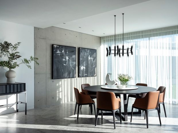 A refined contemporary dining area with a round dark dining table and six upholstered dining chairs, set against a raw concrete feature wall. Floor-to-ceiling sheer curtains provide abundant natural light, while a linear sculptural pendant hangs above the table. Abstract artwork, a sideboard, and a potted plant complete the minimalist interior.