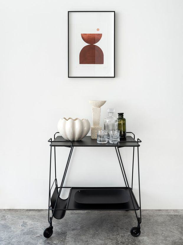 A minimalist styled vignette against a white wall, featuring a black metal two-tier rolling bar cart with a lower tray shelf. The cart displays decorative glassware, bottles, a white sculptural bowl, and a textured vase. Above it hangs framed abstract geometric artwork in earthy terracotta tones. The setting has a grey concrete-look floor and a clean, restrained palette.