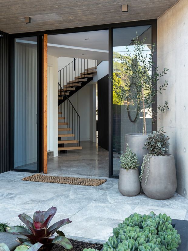 Contemporary residential entry viewed from an outdoor courtyard or terrace, featuring large black-framed glazed doors, a warm timber post, concrete walls, grey stone paving, and sculptural potted plants. The interior beyond contains a minimalist open staircase with timber treads and slender black metal balustrades.