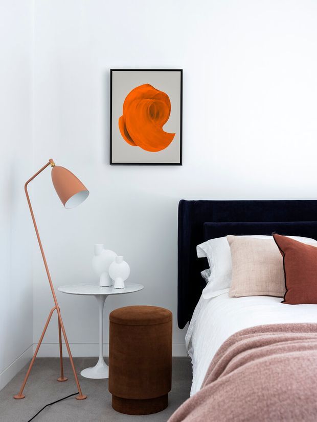A refined bedroom vignette with a navy upholstered bed and white bedding, layered blush and rust cushions, and a soft mauve throw. A slim copper-toned floor lamp stands beside a small white pedestal side table with sculptural white decor, while a brown upholstered cylindrical stool sits in the foreground. The room has pale walls, grey carpet, and framed abstract orange artwork.