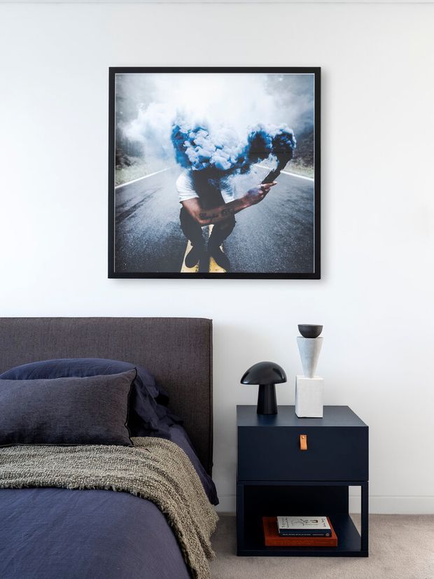 A refined modern bedroom vignette with a dark upholstered headboard, deep navy bedding, a textured neutral throw, and a compact dark bedside table styled with sculptural lamps and a vase. A large framed photographic artwork hangs above the bed, set against a clean white wall and pale carpet.