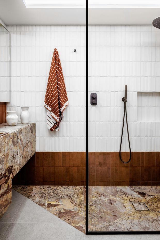 A contemporary bathroom featuring a walk-in shower enclosed by a black-framed glass screen. The walls are clad in vertically textured white tiles with a dark brown tiled wainscot, while the shower floor and vanity side are finished in richly veined natural stone. A wall-mounted bronze shower set, recessed shelf, skylight, and warm rust-coloured towel add detail.