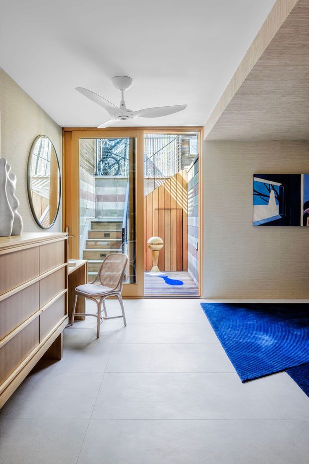 A bright, narrow contemporary entry hall or living area with pale large-format tiled flooring, textured light walls, and warm timber-framed glazing opening to a small courtyard or entry vestibule. A low timber drawer unit with mirror and sculptural decor lines the left wall, while a woven lounge chair, wall-mounted artwork, blue rug, and white ceiling fan add character. The space has strong indoor-outdoor connection and abundant natural light.