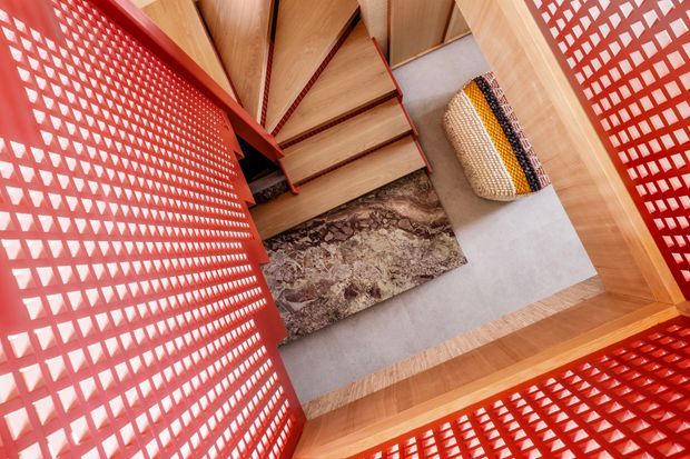 Top-down view of a compact stair landing or entry space framed by vivid red perforated screens. A timber staircase with red trim descends beside a pale grey floor, with a patterned brown rug and woven striped cushion adding texture. Timber framing and ceiling panels create a warm, graphic interior with strong geometric detailing.