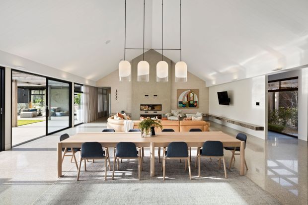 Bright open-plan contemporary living and dining area with a long timber dining table and six dark upholstered chairs in the foreground. Behind it is a lounge with neutral sofas, a large textured feature wall, linear fireplace, wall-mounted television, and sculptural pendant lights. Full-height black-framed glazing opens to an outdoor area, while polished light flooring and a restrained neutral palette create an airy, refined interior.