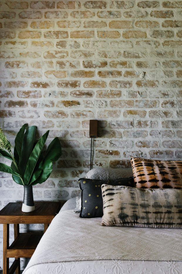 A warmly styled bedroom vignette featuring a textured exposed brick feature wall, layered neutral bedding, patterned cushions, a dark wood bedside table, a leafy plant, and a wall-mounted bedside lamp. The composition has a relaxed, earthy and eclectic character.
