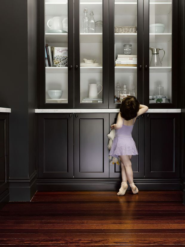 A dark, traditional built-in display cabinet with black painted timber cabinetry, glass-front upper doors, paneled lower doors, and light marble or stone trim. The shelves display white ceramics, glassware, books, and baskets. A young ballerina stands in front of the cabinetry, with polished dark timber flooring below.