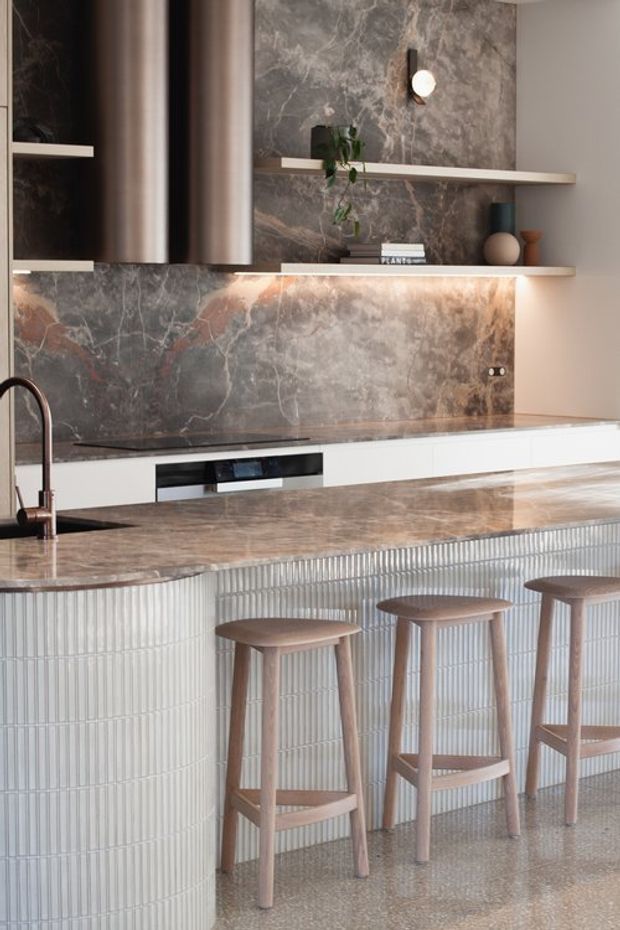 Contemporary kitchen featuring a large island with a vertically ribbed light tile base, a richly veined grey-brown stone benchtop and splashback, white flat-panel cabinetry, dark cylindrical rangehoods, open display shelving, and warm timber bar stools. Integrated lighting under the shelving adds a soft glow to the refined, material-focused space.