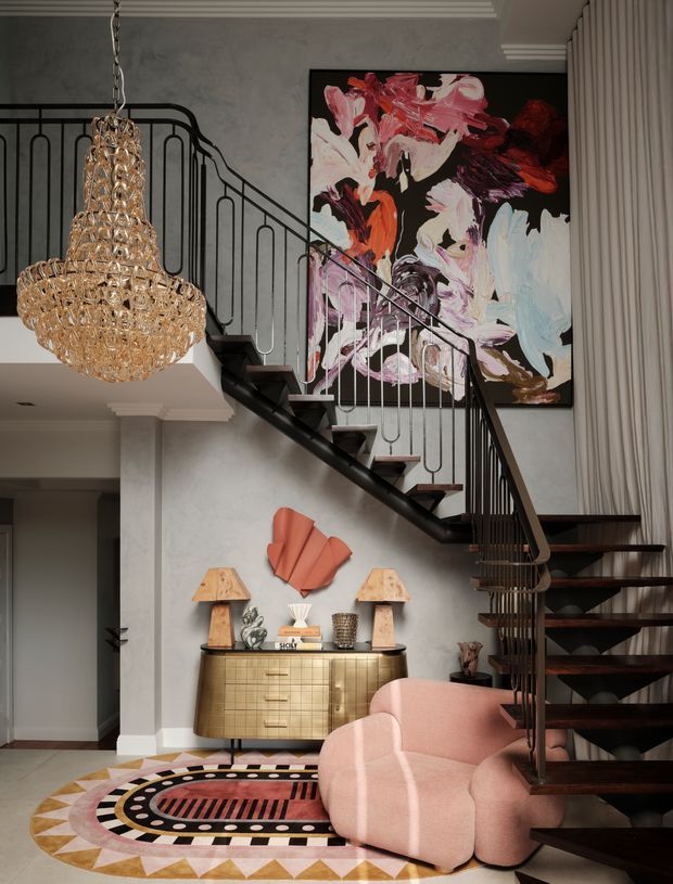 A sophisticated interior foyer beneath a staircase, featuring a sculptural black metal stair balustrade, dark timber stair treads, a large abstract artwork, a gold-toned sideboard, pink curved lounge seating, a patterned geometric rug, and an ornate crystal chandelier. The space has layered decorative detailing, rich contrast, and an art-led, glamorous atmosphere.