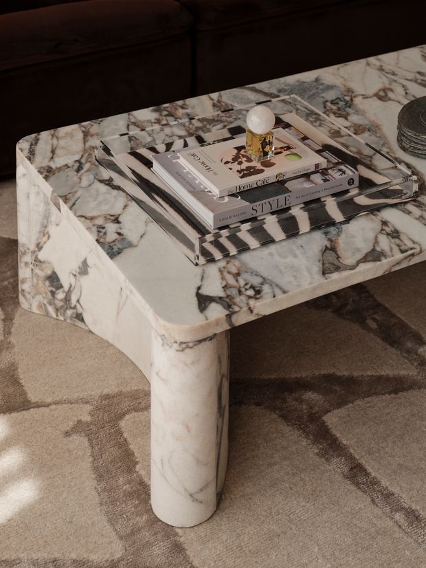 Close-up interior styling photograph of a low, rounded rectangular coffee table with a thick veined marble top and matching cylindrical marble legs. The table sits on a neutral taupe geometric rug in front of a dark upholstered sofa. Stacked design books, a striped tray, and a small decorative object create a styled tabletop arrangement. The image has a warm, refined, luxury-oriented palette and subdued lighting.