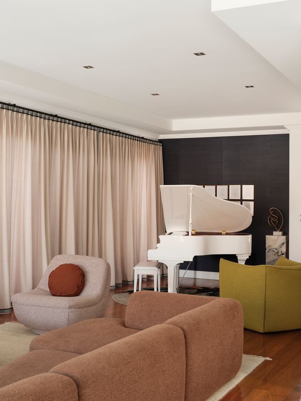 A refined living room with a white grand piano positioned against a dark feature wall, framed artwork, and sculptural decor. Full-height blush-beige curtains span one side of the room, while rounded upholstered seating in soft beige, terracotta, brown, and olive green surrounds a light rug. The room has recessed ceiling lighting, timber flooring, and a warm, softly layered contemporary interior.