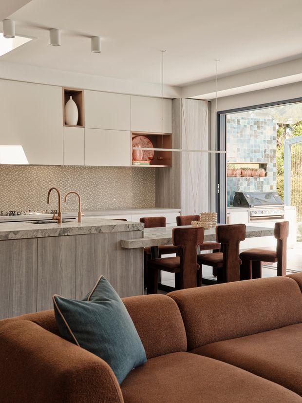 A contemporary open-plan kitchen and dining area viewed from a living room. The kitchen features pale grey wood-grain cabinetry, a stone island with an integrated sink and copper-toned taps, patterned tile splashback, and open display shelving. A dining table with upholstered brown chairs sits beside the island, while a large glazed door opens to an outdoor entertaining area with a built-in barbecue. A brown sectional sofa with a teal cushion fills the foreground. The space has warm neutral finishes, layered lighting, and strong indoor-outdoor connection.